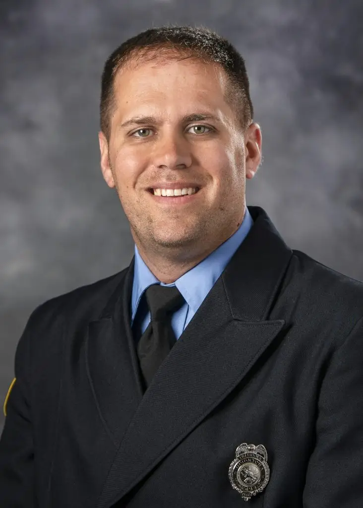 A man wearing a dark blue uniform with an emblem on the left side of his chest is smiling at the camera. He has short hair and a light complexion. The background is a gray, textured backdrop.
