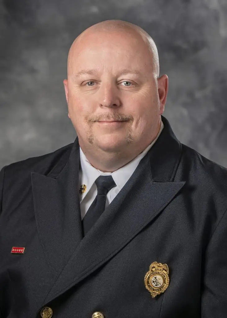 A bald man with a mustache is wearing a formal dark navy uniform with a white shirt and black tie. He has two gold pins on his jacket, one resembling a badge. The background is gray and indistinct.