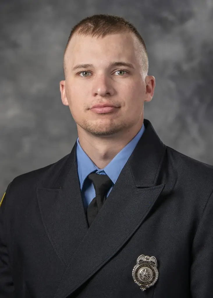 A uniformed male firefighter, wearing a dark formal jacket with a badge on the left side, a light blue shirt, and a black tie, poses for a professional portrait against a gray backdrop. He has short hair and a neutral expression.