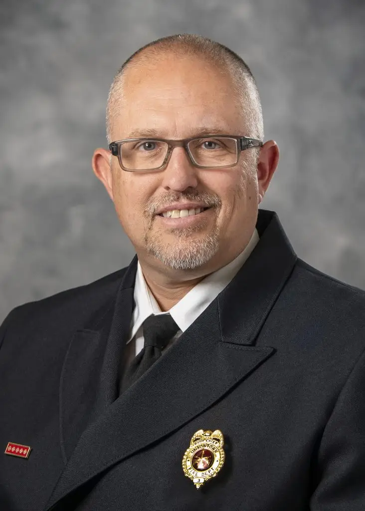 A man with short-cropped hair and glasses is wearing a dark uniform with a badge on the lapel and a tie. He is smiling against a gray, slightly blurred background.