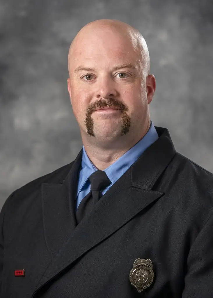 A bald man with a brown mustache is wearing a formal dark uniform with a blue shirt and black tie. A badge is visible on his coat. The background is a gradient of gray. His expression is neutral.