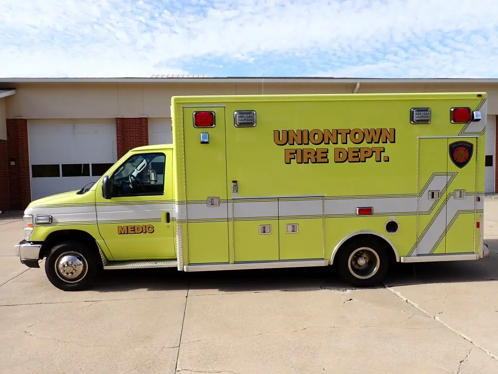 A light green and white ambulance marked "Uniontown Fire Dept." and "Medic 1" is parked outside a building with multiple garage bays. The vehicle features emergency lights on the top and sides, along with reflective striping and insignia.
