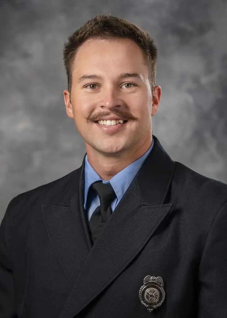 A man with short brown hair and a mustache is smiling at the camera. He is wearing a dark formal uniform with a light blue shirt and a black tie. A silver badge is pinned to his jacket. The background is a gray, blurred texture.