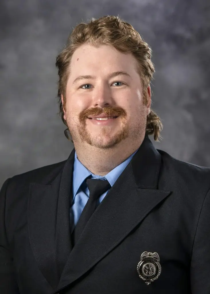 A man with light brown hair and a mustache smiles at the camera. He is wearing a dark blazer with a tie and a light blue shirt. A badge is pinned to his blazer. The background is a gradient of gray and black.