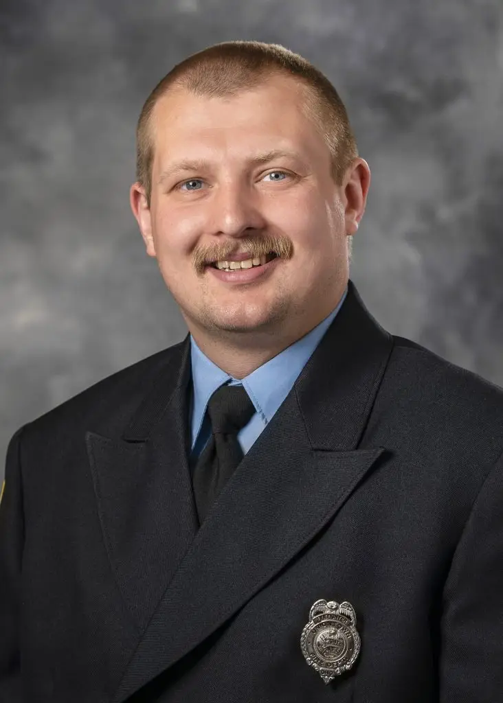 A man with short hair and a mustache is smiling while wearing a dark-colored uniform jacket with a light blue shirt and a black tie. The jacket features a silver badge on the left side. The background is a neutral gray.