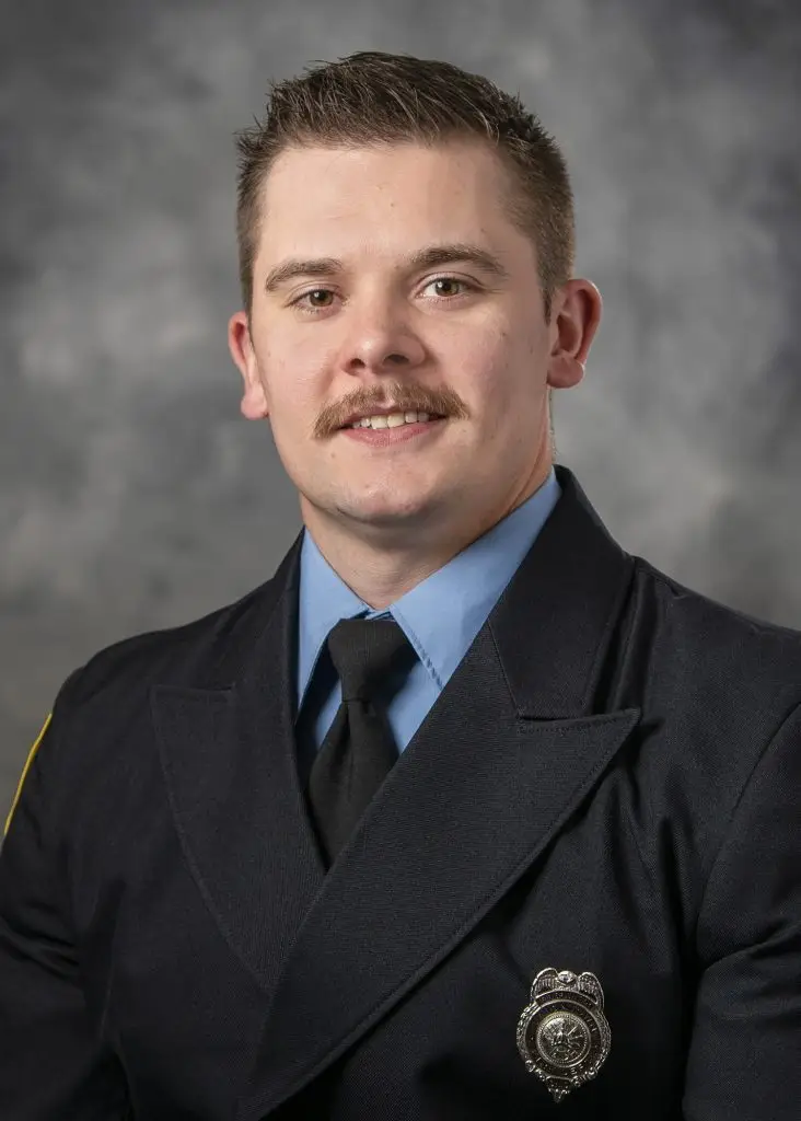 A man with short, light brown hair and a moustache is wearing a formal dark blue uniform with a light blue shirt and black tie. A badge is pinned to his jacket. The background is a blurred, grey backdrop.