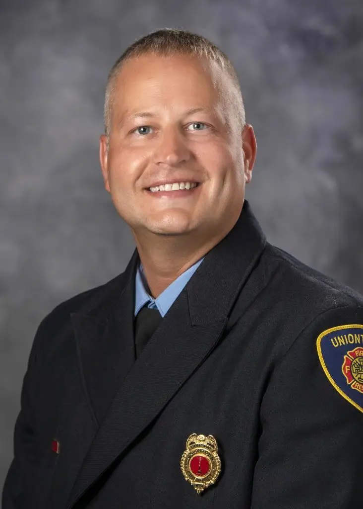 A smiling person with short blonde hair is wearing a dark uniform with a badge and a patch on the shoulder that reads "Union Fire Dept." in gold letters. The background is a gray, cloudy, studio-style backdrop.