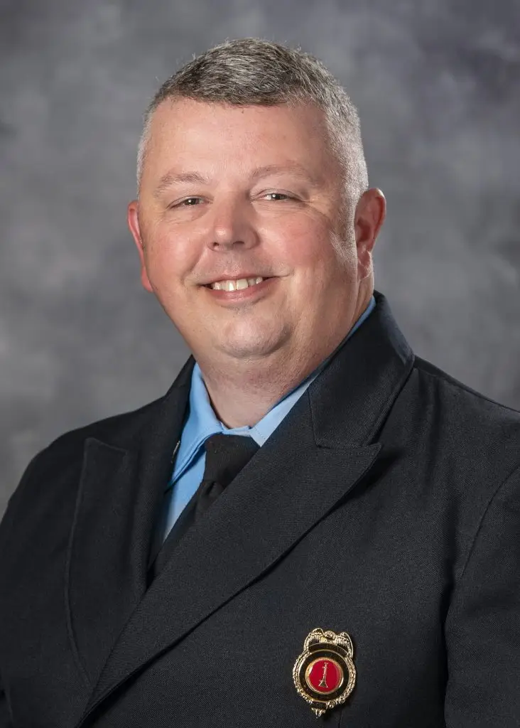 A man with short gray hair is smiling while wearing a dark suit jacket adorned with a red and gold badge on the lapel. He is posing against a gray mottled background.