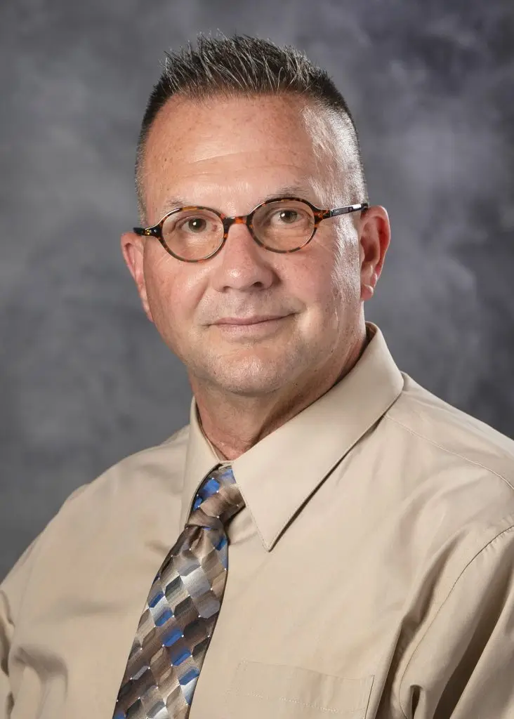 A man with short gray hair, wearing round glasses, a light beige dress shirt, and a multicolored striped tie, poses for a portrait. The background is a gray, slightly textured backdrop.