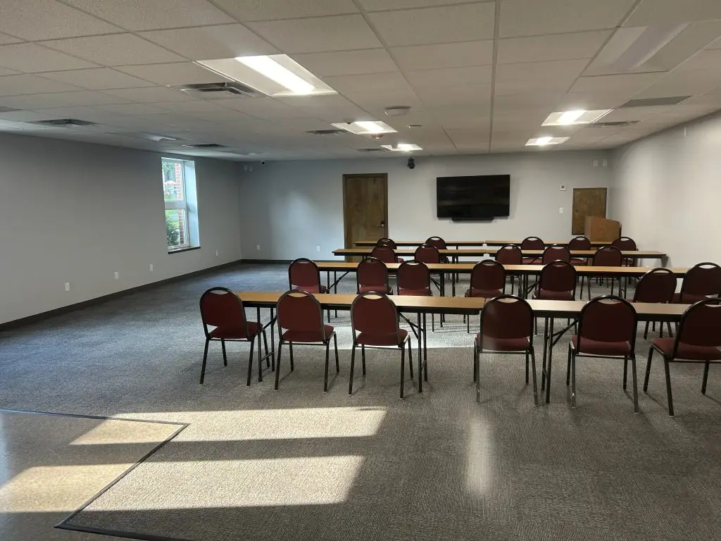 A conference room with several rows of red chairs and wooden tables facing a large flat-screen TV mounted on the wall. The room is well-lit with natural light coming through a window on the left and has white walls with a gray carpeted floor.