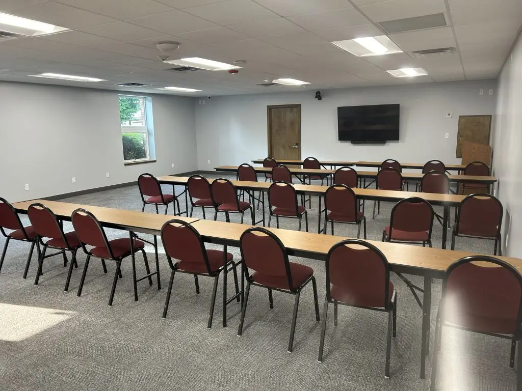 A rectangular conference room with a carpeted floor. Three long tables with black legs are arranged in rows, with red padded chairs on each side. A window on the left wall and a flat-screen TV are mounted on the front wall. The room is empty and well-lit.