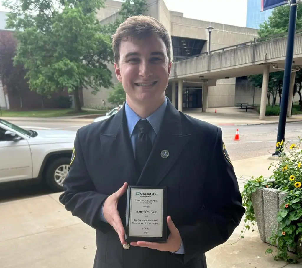 A person in a dark uniform is smiling while holding a plaque in an outdoor setting. The background includes a building, greenery, and a parked white vehicle. The plaque appears to be an award from the Cleveland Clinic.