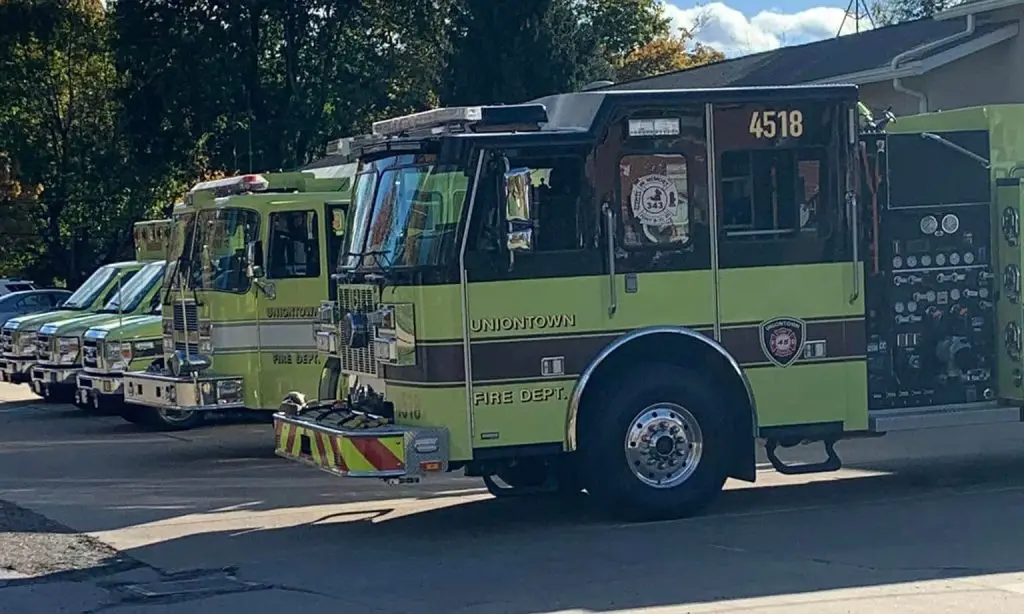 A row of three green fire trucks is parked in front of a fire station. The closest truck, marked with "4518" and "Uniontown Fire Dept," partially obscures the other two. Various equipment and control panels are visible on the trucks. Trees and a building are in the background.
