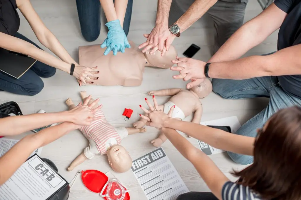 A group of people practice CPR on adult and infant mannequins, pressing down on their chests. Training materials, including a First Aid booklet and CPR instructions, are scattered on the floor. One participant wears blue medical gloves.