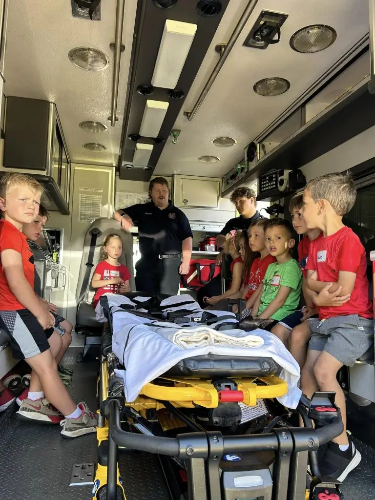 Several children and a paramedic are inside an ambulance. The paramedic is explaining something and gesturing with his hands. The children are seated on the benches on either side, listening attentively. A stretcher is in the center of the vehicle.