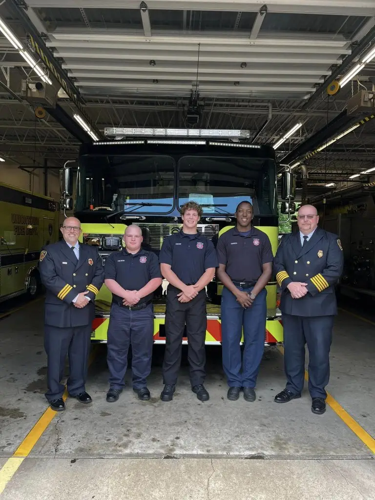 A group of five firefighters stand in front of a fire truck inside a fire station. Two are in uniform, while the other three are in formal dress uniforms. They are posing for a photo and smiling. The fire station's garage and equipment are visible in the background.