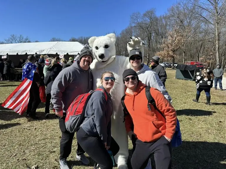 Four smiling people pose outdoors with someone in a polar bear costume. The group is in a grassy area with other participants, a tent, and bare trees visible in the background. It appears to be a chilly day, as everyone is dressed warmly.