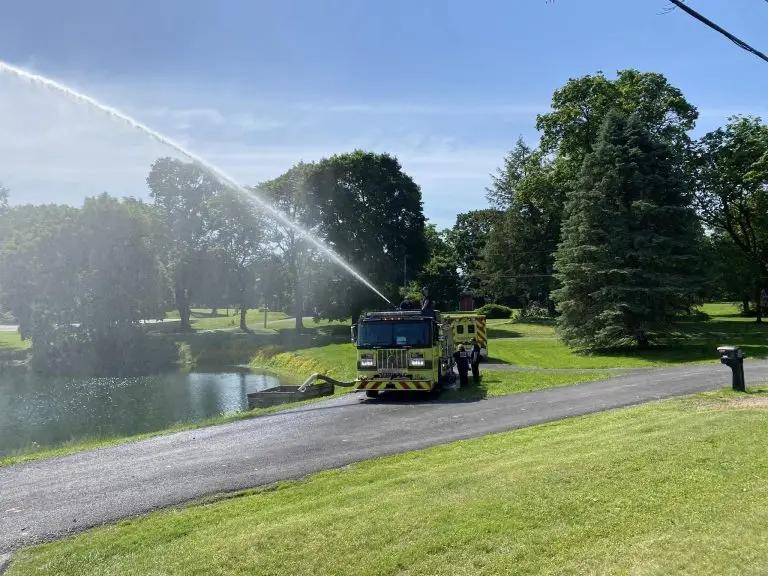 A fire truck spraying water into a pond is parked on a paved road amidst a grassy area with trees and a mailbox visible. The sky is clear and blue with sunlight illuminating the scene.

.