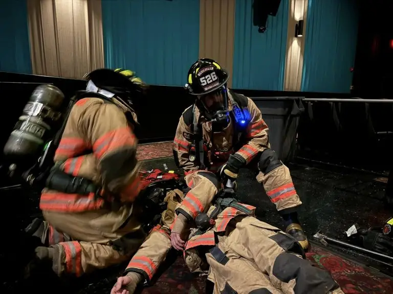 Two firefighters in full gear, kneeling and attending to a colleague lying on the ground indoors. One is checking the colleague's condition while the other appears to be preparing equipment. The scene is inside a dimly lit room with blue and beige curtains.