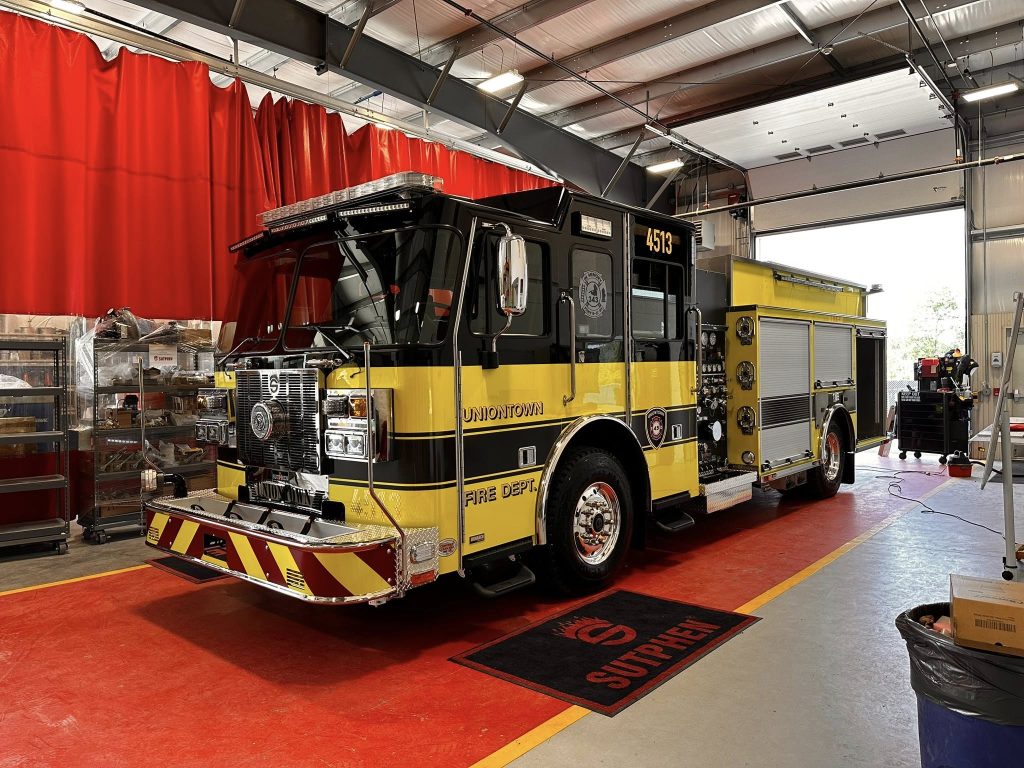 A black and yellow fire truck marked "Uniontown Fire Dept." is parked inside a garage with red flooring and red curtains. Shelves with equipment and tools are visible in the background.